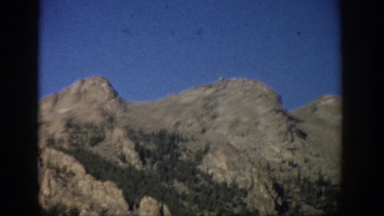 1959 Colorado 8mm frame showing rugged mountain peaks, sparse evergreens, and blue sky within dark film or window-like borders.