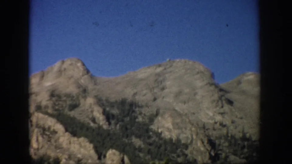 1959 Colorado 8mm frame showing rugged mountain peaks, sparse evergreens, and blue sky within dark film or window-like borders.