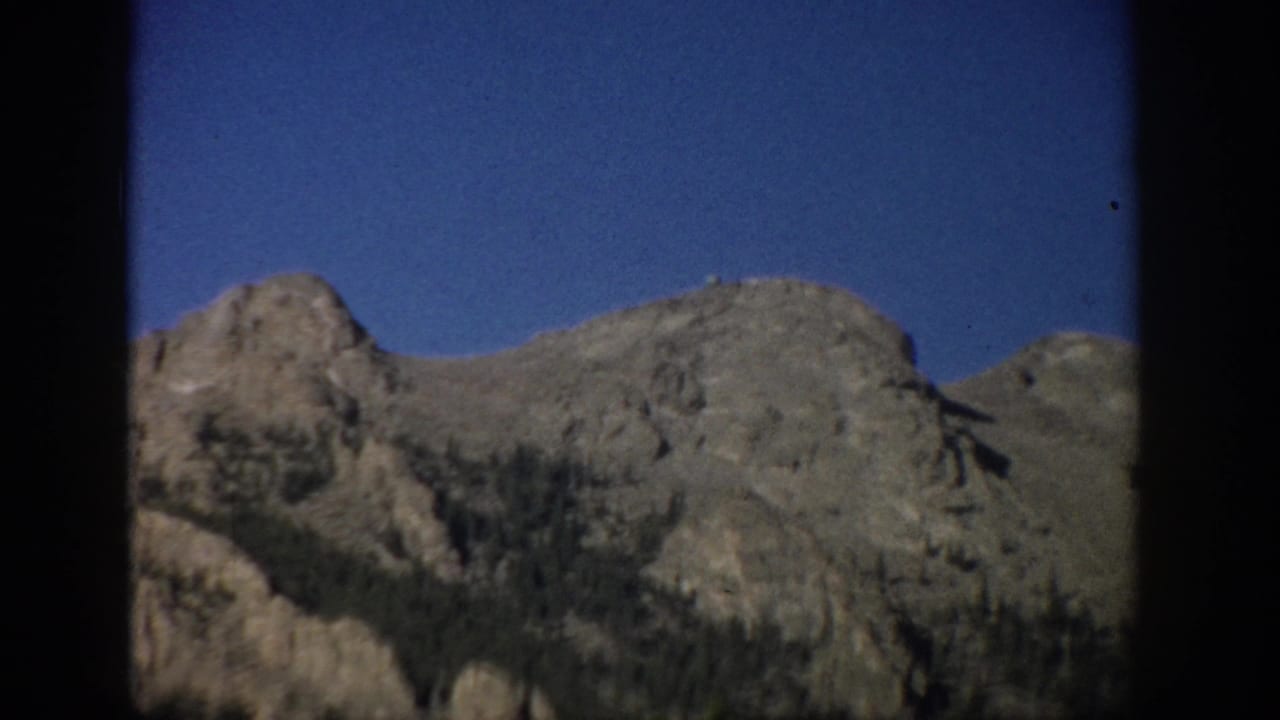Mountain peaks and tree-covered lower slopes beneath a clear sky, framed by dark vertical edges.