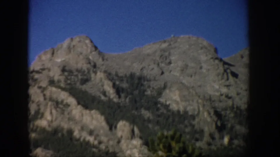 Colorado 1959 home-movie frame showing a rugged mountain ridge, rocky outcrops, evergreens, and clear sky through dark borders.