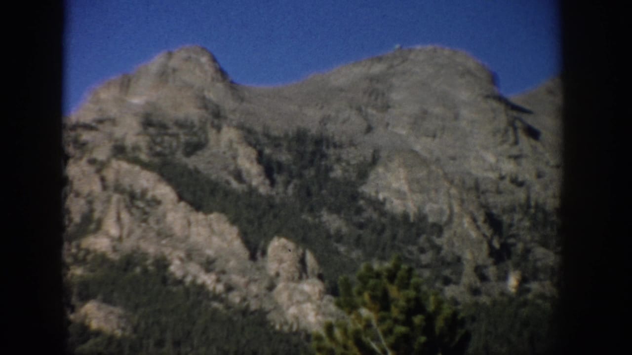 1959 Colorado home-movie frame showing gray rocky mountain terrain above dark evergreen forest and blue sky.