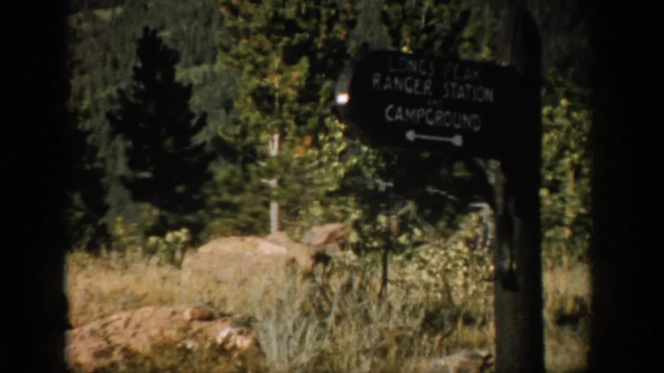1959 Colorado frame showing a directional sign for Longs Peak Ranger Station and campground amid trees, grass, and boulders.