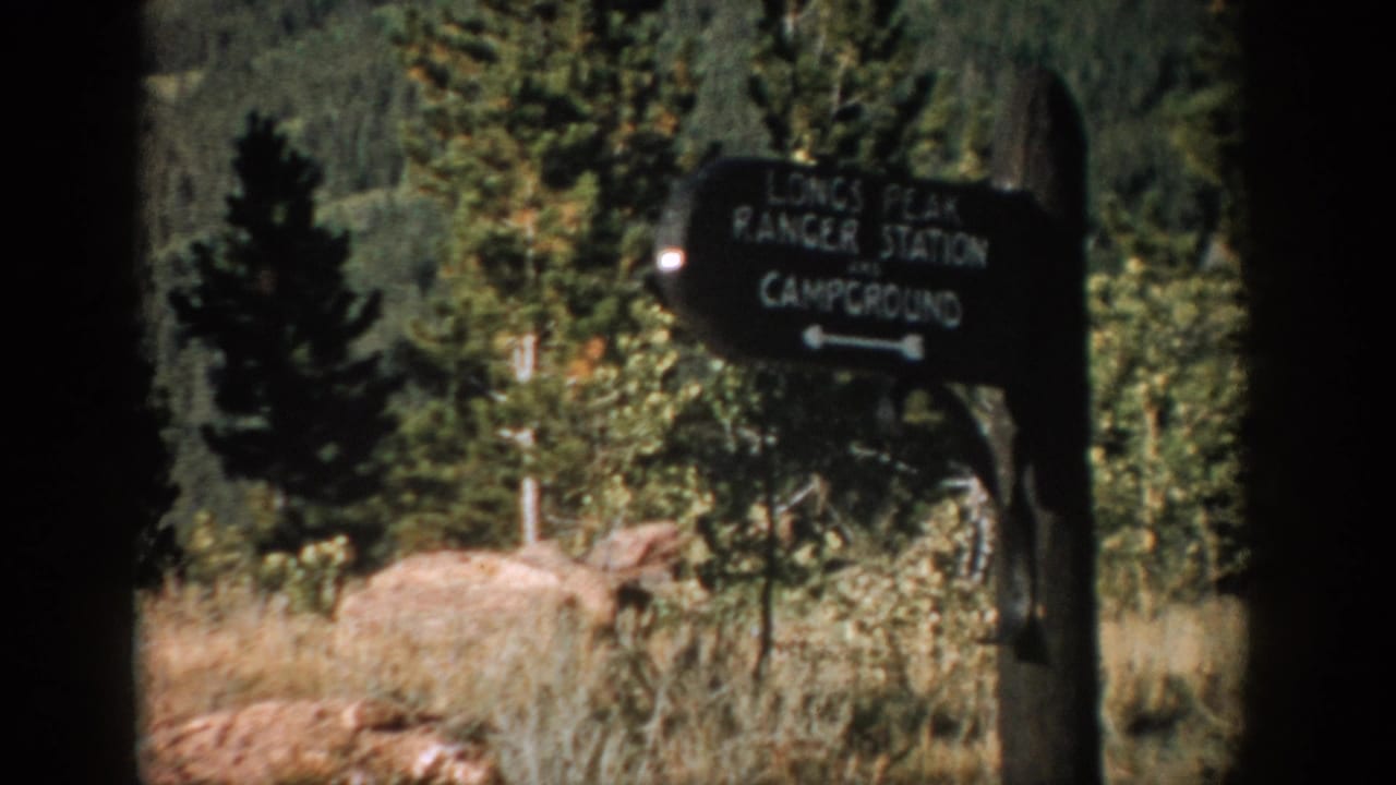 Longs Peak Ranger Station sign beside trees, grass, and pale rocks.
