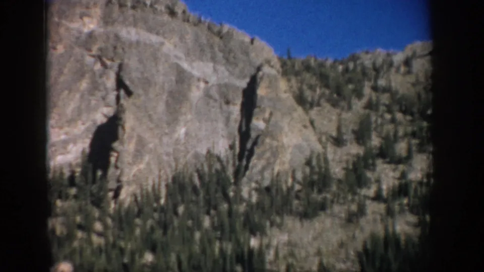 1959 Colorado 8mm frame showing a rugged mountain rock face, tree-covered lower slope, and clear blue sky.