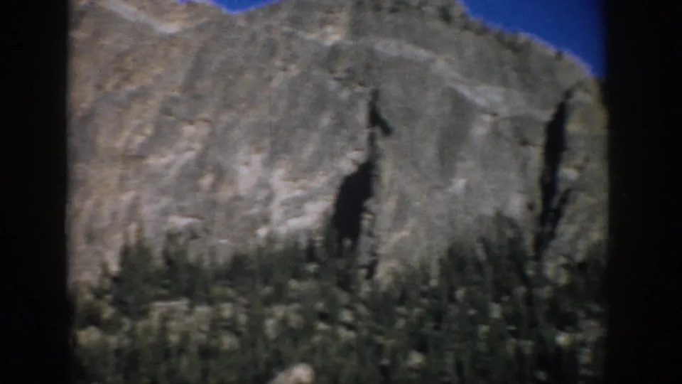 1959 Colorado home-movie frame of a gray rock formation and forested lower slope under a blue sky.