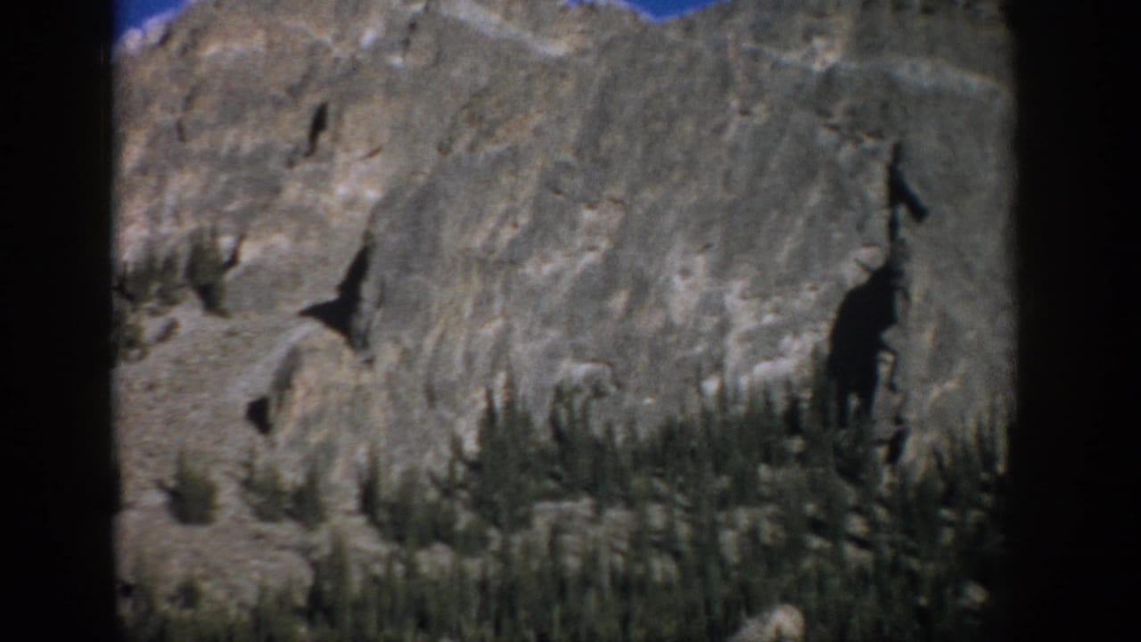 1959 Colorado home-movie view of a gray cliff face with scattered evergreens, loose rock, blue sky, and soft 8mm film edges.