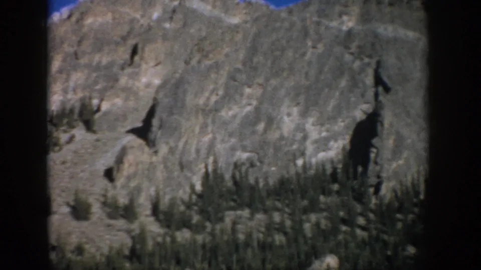 1959 Colorado home-movie view of a gray cliff face with scattered evergreens, loose rock, blue sky, and soft 8mm film edges.