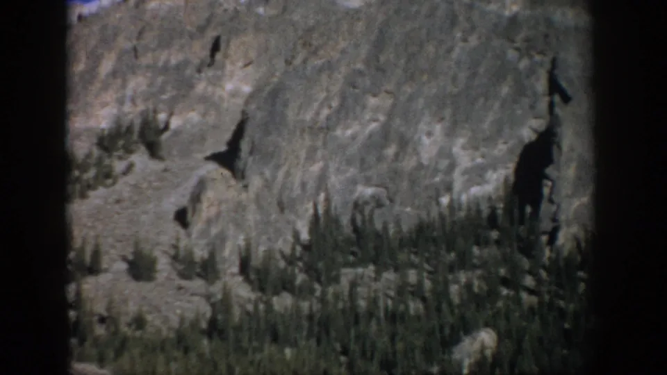 1959 Colorado 8mm frame showing rugged rock formations, conifer trees on a steep slope, and a narrow strip of blue sky.