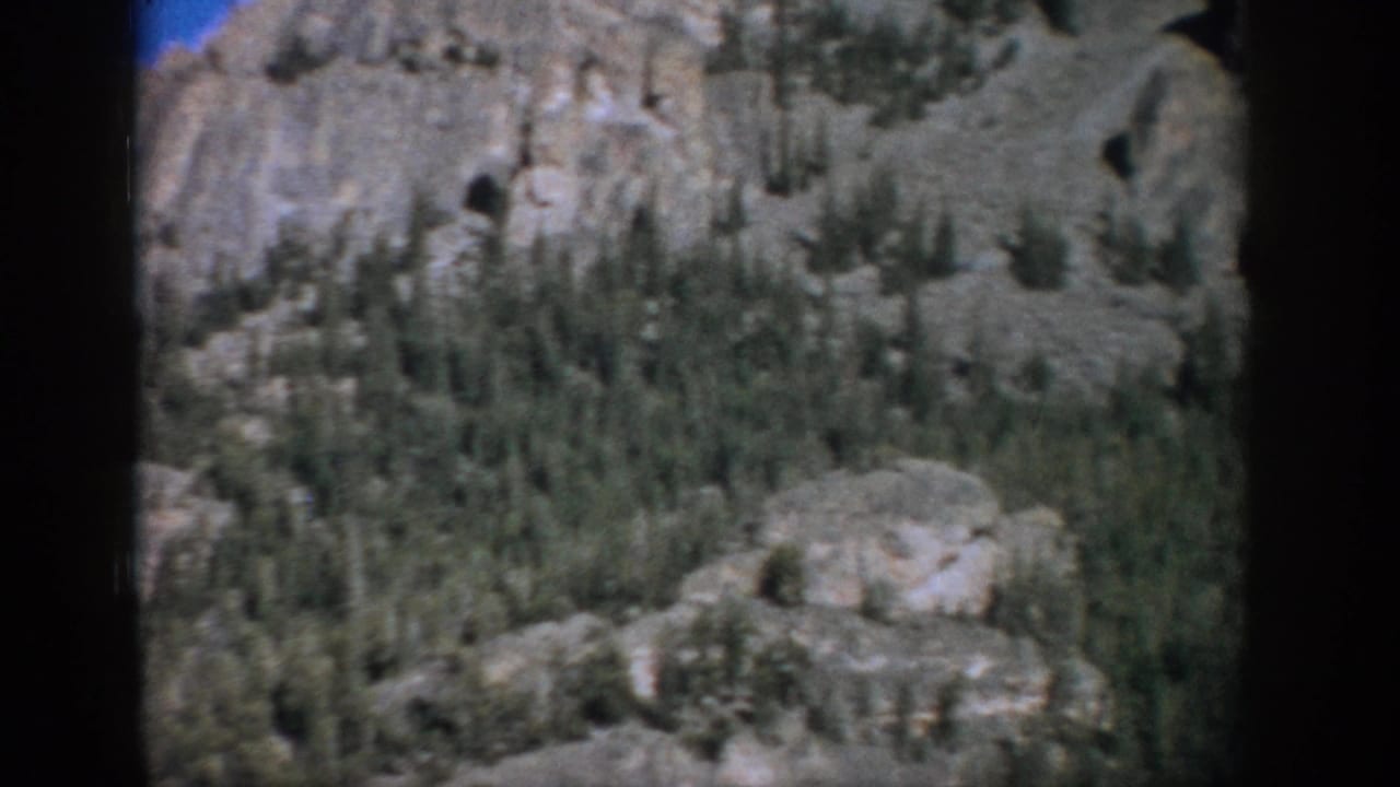 Rocky mountain slope with evergreen trees beneath a blue sky, framed by dark film edges.