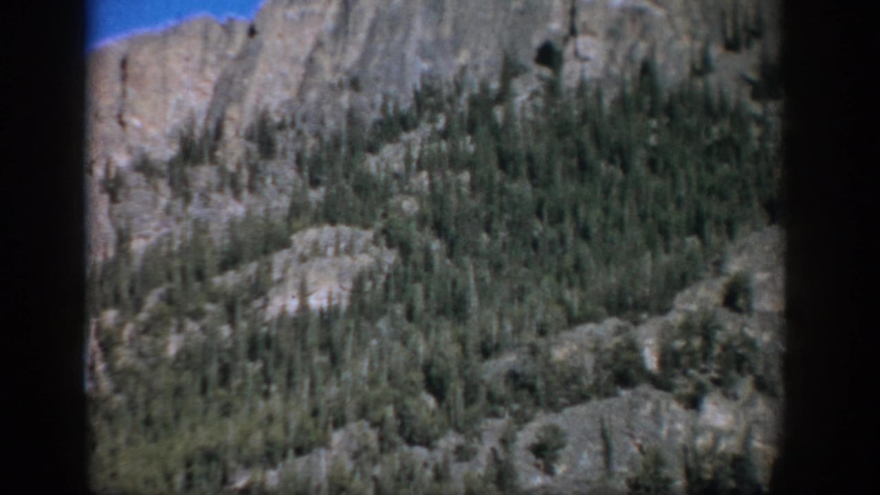 1959 Colorado home-movie frame of a rugged, forested mountain slope with pale rock outcrops and 8mm frame edges.