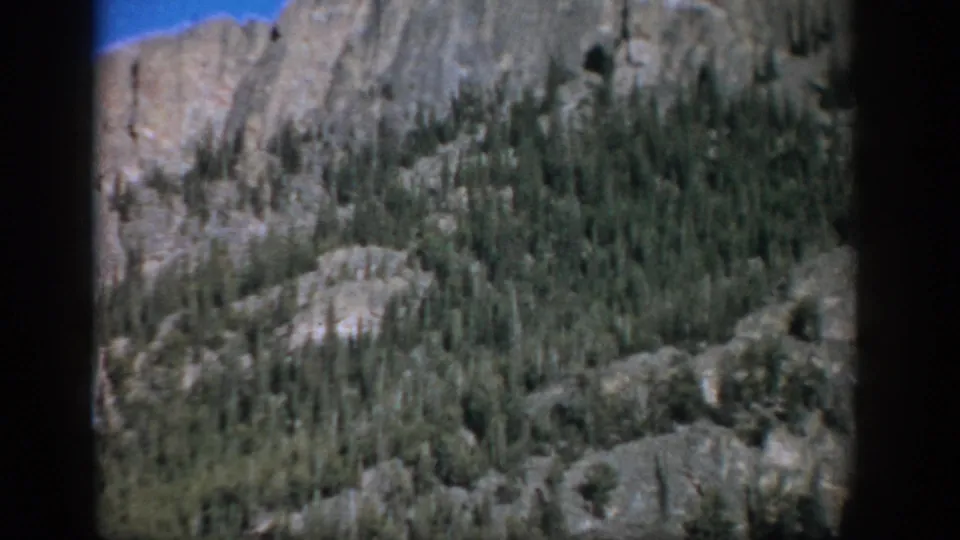 1959 Colorado home-movie frame of a rugged, forested mountain slope with pale rock outcrops and 8mm frame edges.