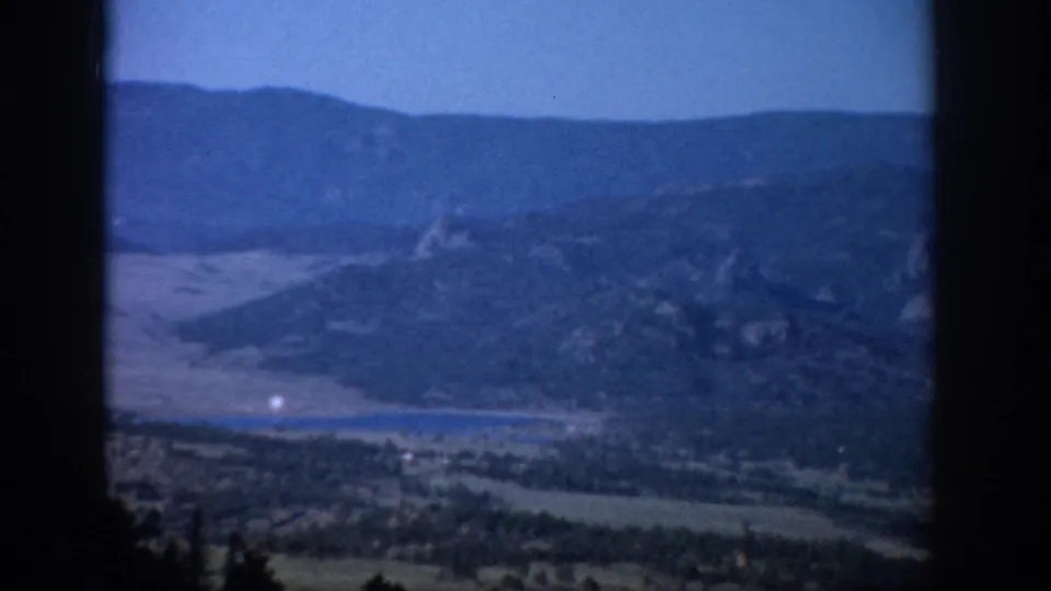 1959 Colorado 8mm vista showing distant mountains, a lake or reservoir, valley floor, and silhouetted foreground trees.