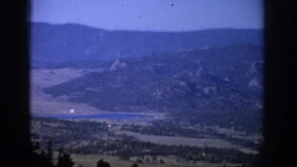 Blue-shifted 1959 Colorado home-movie frame with distant mountain ridges, valley terrain, water, and foreground tree silhouettes.