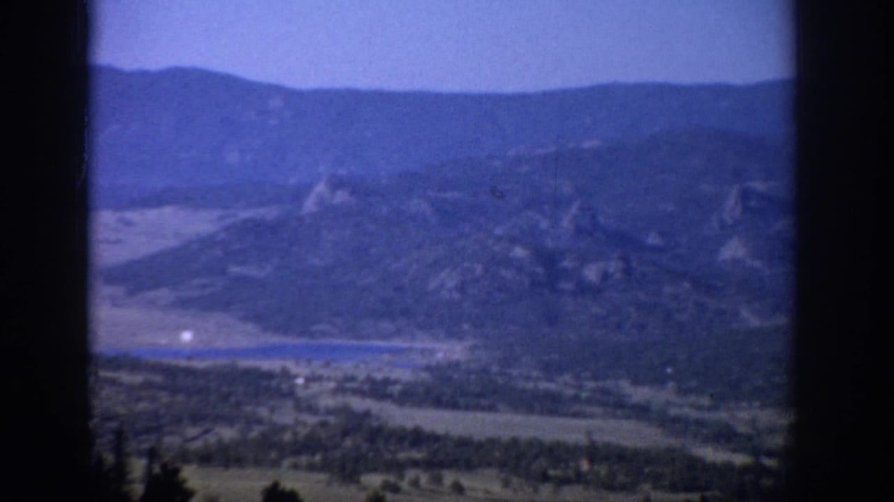 1959 Colorado 8mm frame looking through a dark window frame toward mountains, a green valley, and a small lake.