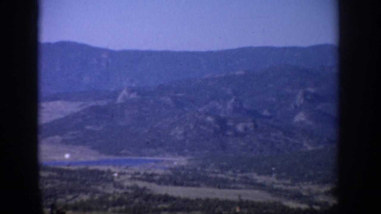 1959 Colorado home-movie frame with blue-cast mountains, valley fields, and a lake divided by a window mullion.