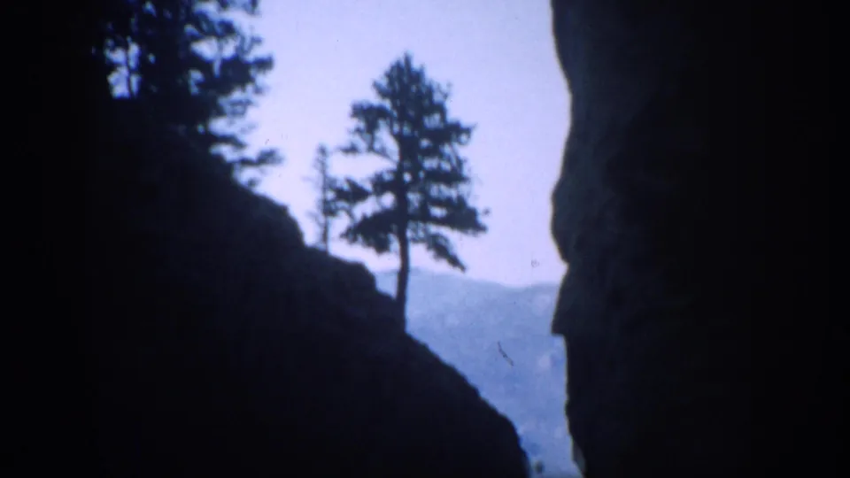 1959 Colorado 8mm frame showing dark rock walls and pine silhouettes with distant mountains beyond the canyon opening.