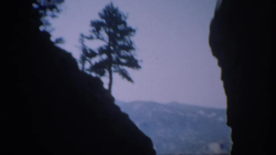 Colorado canyon view from moving 8mm footage, with a pine tree silhouetted above dark rock and a hazy mountain ridge in back.