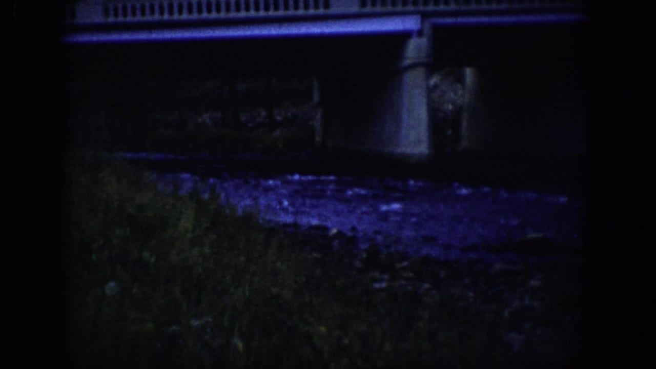 1959 Colorado 8mm frame with a concrete bridge railing and support above a dim riverbank and purple-blue reflected water.