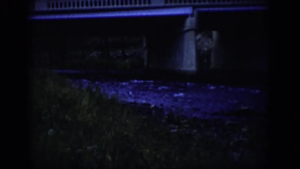 1959 Colorado 8mm frame with a concrete bridge railing and support above a dim riverbank and purple-blue reflected water.