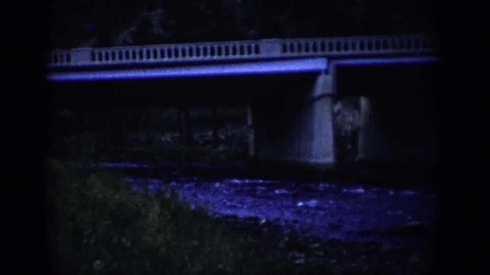 1959 Colorado 8mm frame showing the bridge more clearly, with railing, support column, dark river surface, and grassy foreground.