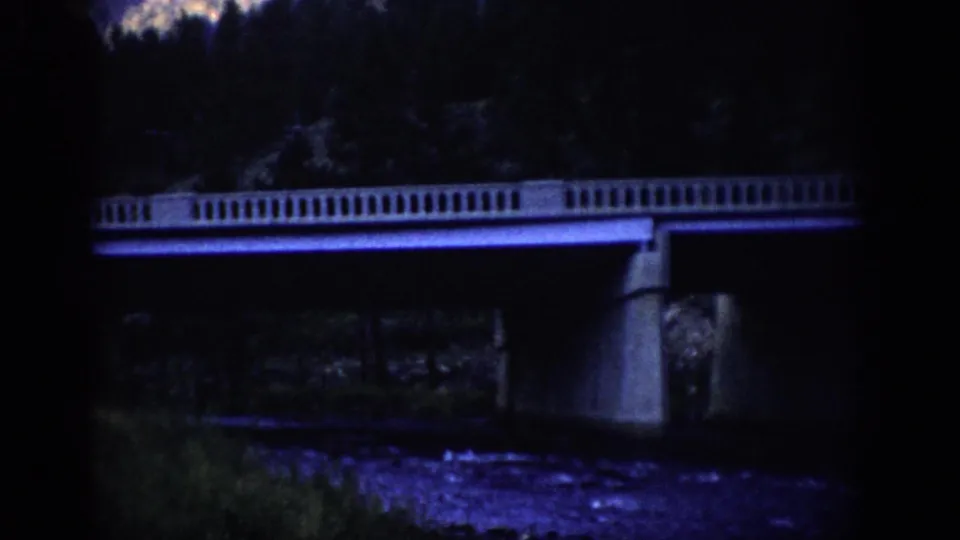 1959 Colorado 8mm frame of a concrete bridge over dark water, with riverbank vegetation and trees barely visible in blue-purple light.