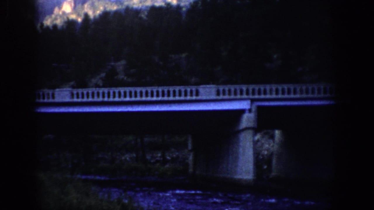 1959 Colorado 8mm frame showing a concrete bridge with balustrade above a river, backed by evergreen slopes and rocky peaks.