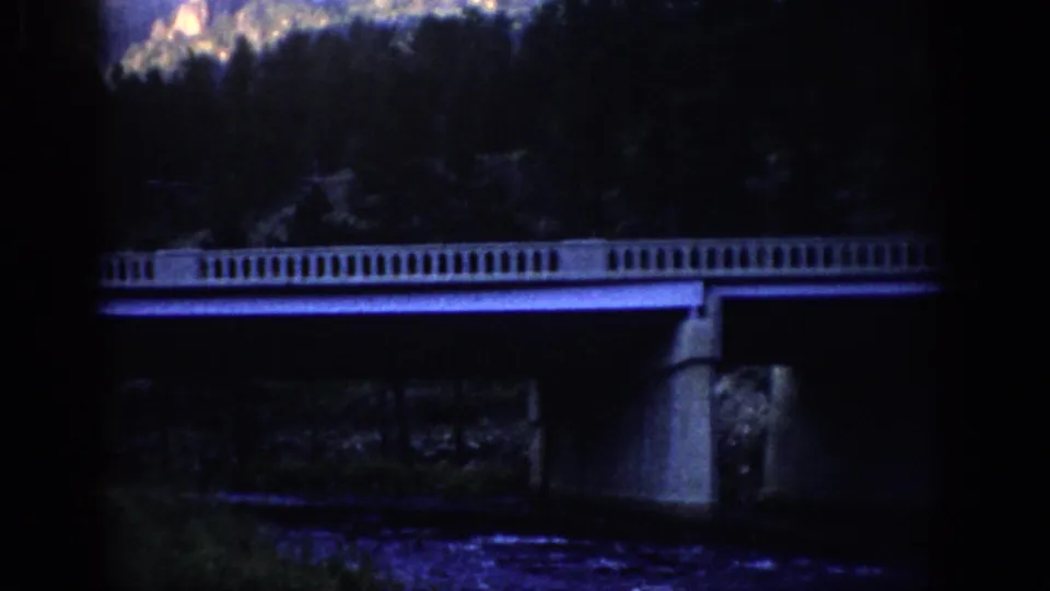 1959 Colorado 8mm frame showing a concrete bridge with balustrade above a river, backed by evergreen slopes and rocky peaks.