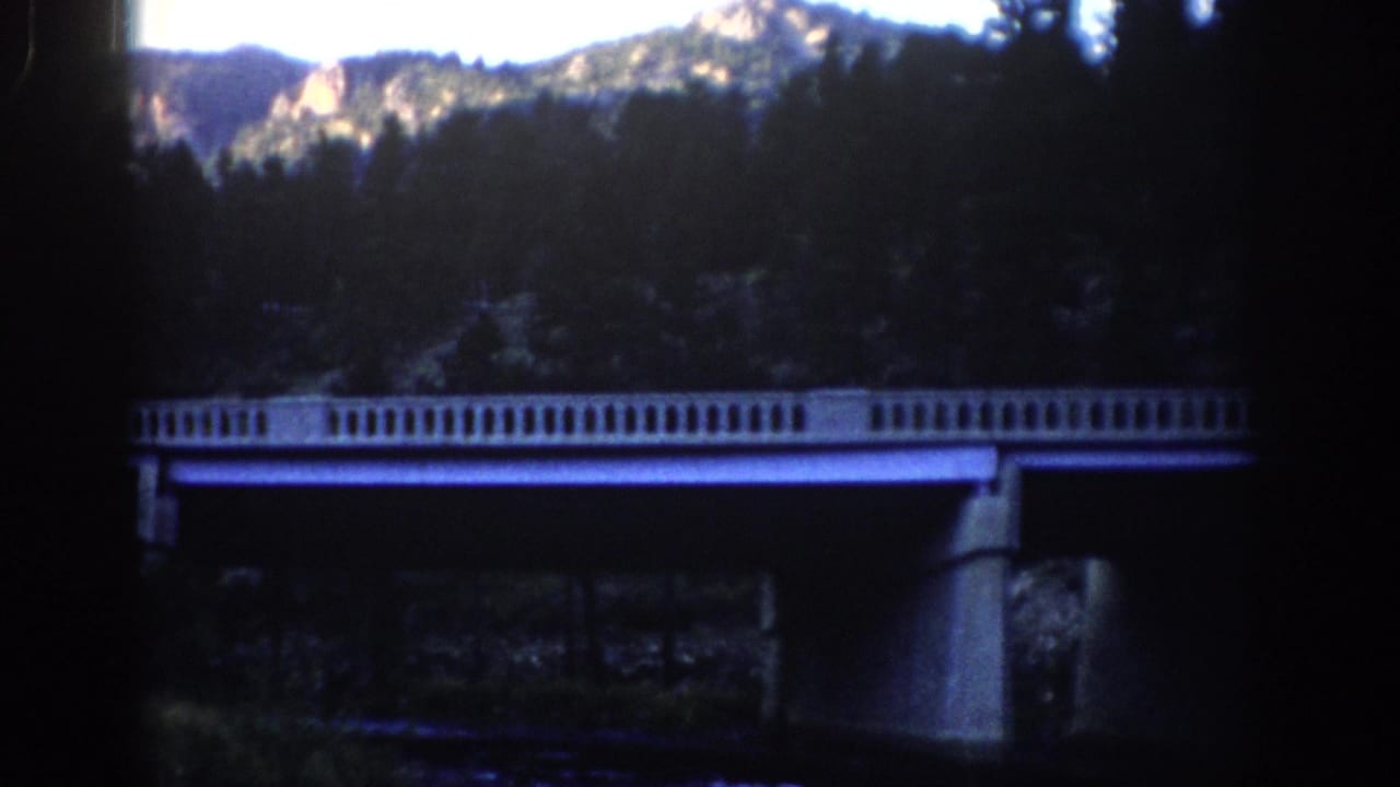 Bridge and forested mountain slope with bright rocky peaks beyond.