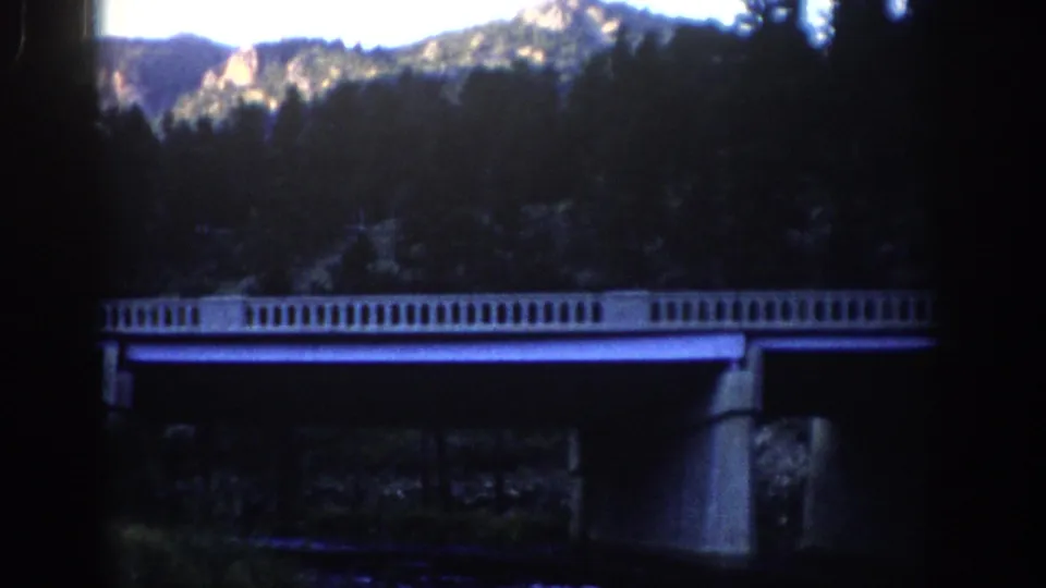 1959 Colorado home-movie frame with a pale concrete bridge set against dark trees and sunlit mountain rock.