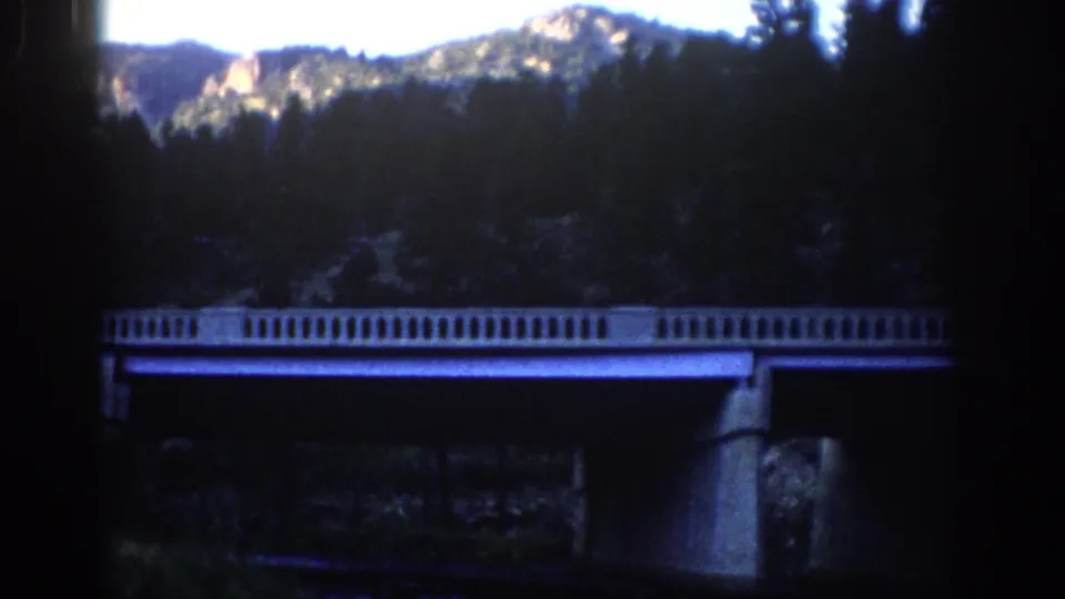 1959 Colorado 8mm still showing a bridge over a dark river area, with trees and rugged peaks in the background.