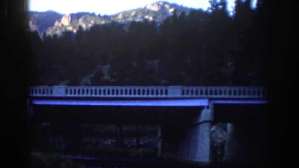 Final 1959 Colorado frame holds on the concrete bridge, dark trees, and sunlit rocky mountain backdrop in aged 8mm color.