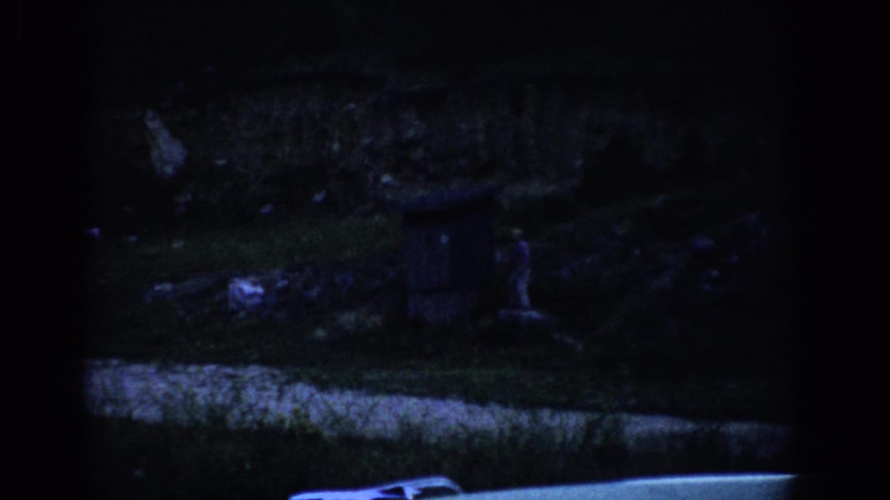 1959 Colorado 8mm frame showing a shadowed grassy hillside, a small distant figure, a dark box-like structure, and a vehicle edge below.