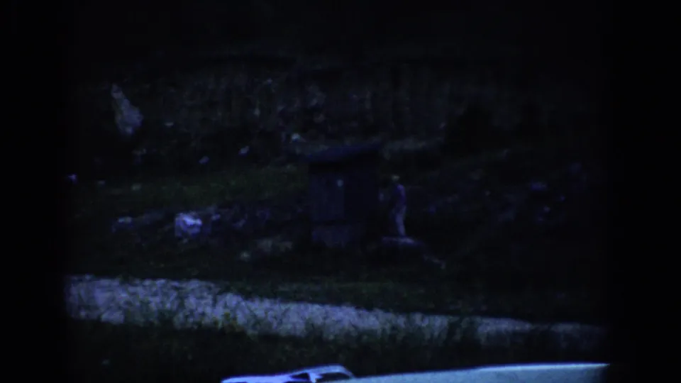 1959 Colorado 8mm frame showing a shadowed grassy hillside, a small distant figure, a dark box-like structure, and a vehicle edge below.