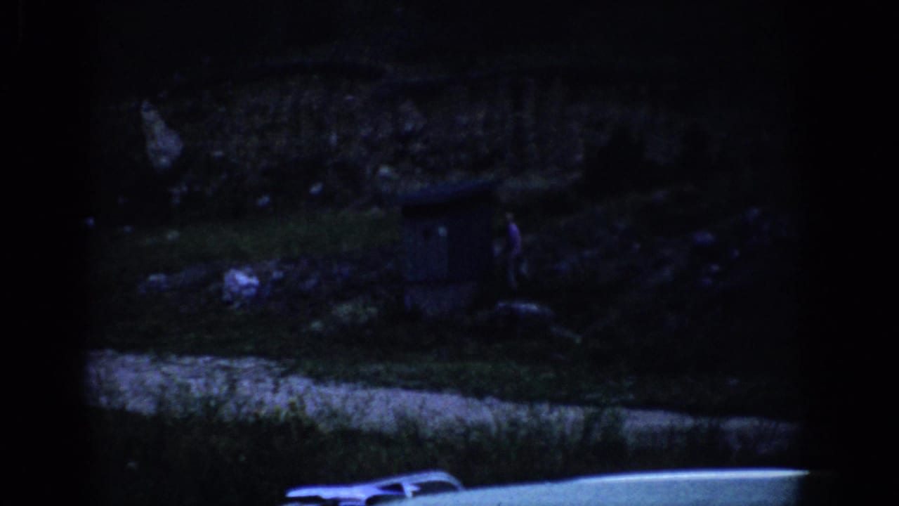 Shadowed 1959 Colorado home-movie frame with a small figure on the hillside, a dark structure, scattered ground detail, and road edge below.