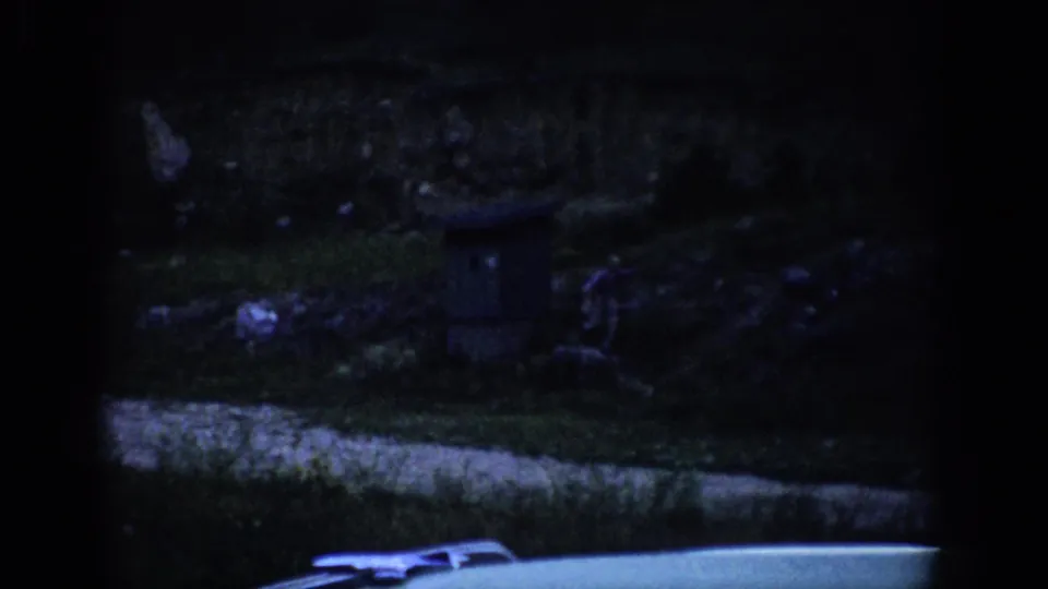 Low-light 8mm frame from Colorado, 1959, showing a small distant figure on a dark grassy slope above a bright road edge.