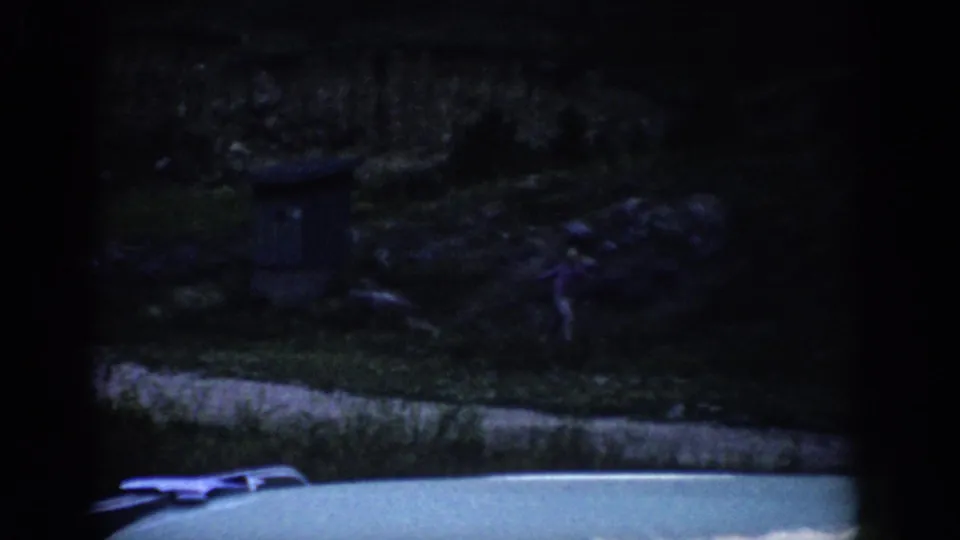 1959 Colorado 8mm frame with a small distant figure near the right edge of a shadowed hillside above a pale roadside foreground.