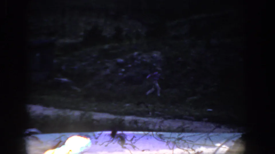 1959 Colorado home-movie frame showing a dark shoreline or bank beside lake water, with soft exposure and visible 8mm frame edges.