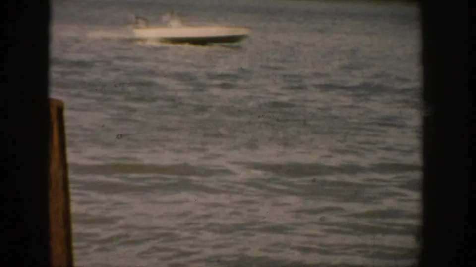 A small motorboat crosses a Colorado lake in distant 1959 8mm footage, with rippled water and dark home-movie borders.