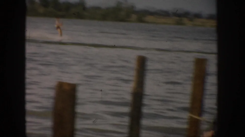 A distant person appears to water ski on a Colorado lake in 1959, with wooden posts and shoreline visible in the foreground and background.