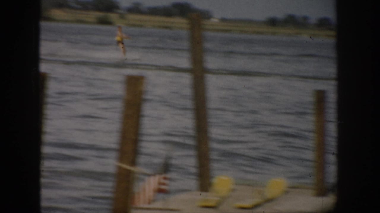 1959 Colorado 8mm frame with dock posts, a small American flag, and a distant water skier crossing calm lake water.