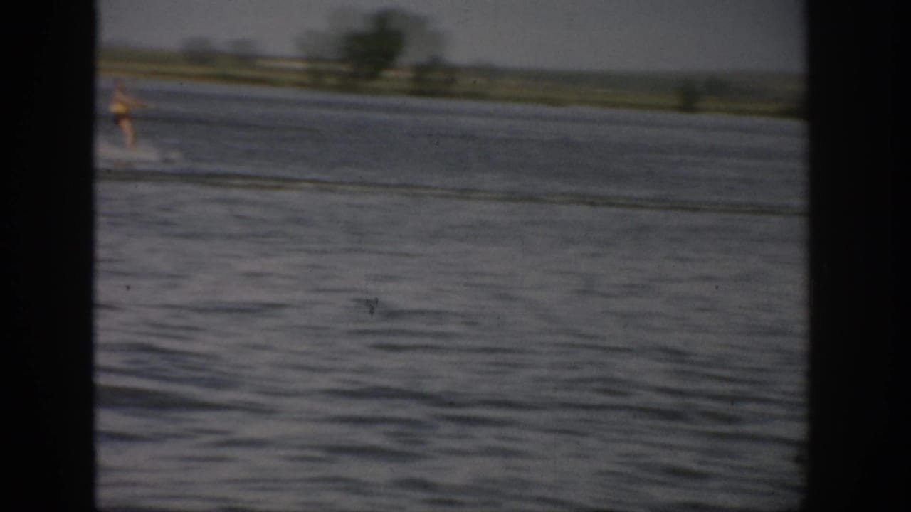 1959 Colorado 8mm frame showing a small, distant water skier moving across the lake near a low rural shoreline.
