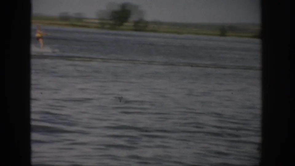 1959 Colorado 8mm frame showing a small, distant water skier moving across the lake near a low rural shoreline.