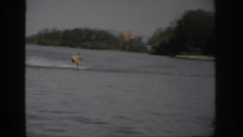 1959 Colorado 8mm frame showing a distant water skier and white wake against a broad lake and tree-lined shoreline.