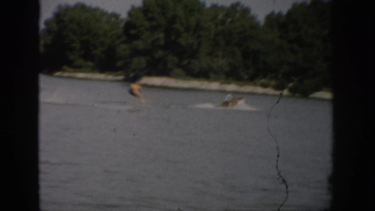 1959 Colorado home-movie frame showing a water skier crossing the lake behind a boat wake, seen through dark foreground framing.