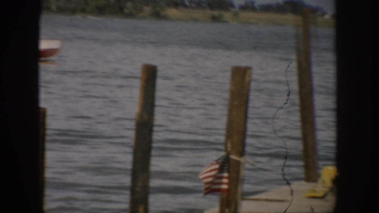 1959 Colorado lake view with wooden dock posts, an American flag, and water beyond, framed by dark edges from the filming position.