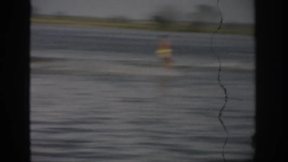 A blurred 1959 Colorado water-skier figure crosses open lake water, emphasizing motion and home-movie camera softness.