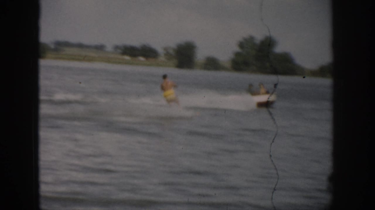 1959 Colorado home-movie frame showing a distant water skier in yellow trunks pulled across a lake by a small boat.