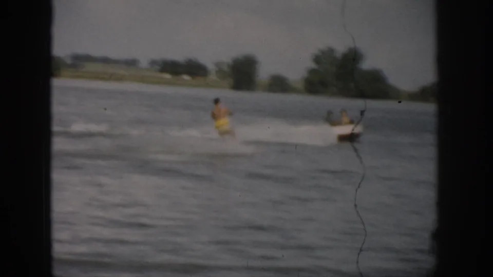 1959 Colorado home-movie frame showing a distant water skier in yellow trunks pulled across a lake by a small boat.
