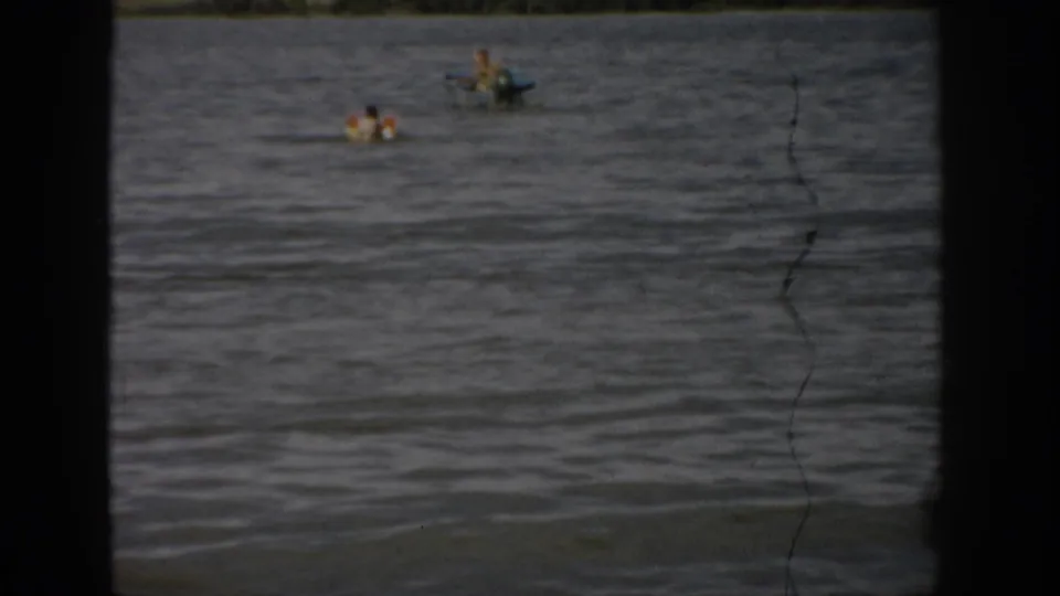 Home-movie frame from the Colorado lake scene showing several distant people in the water and a small boat on choppy lake surface.