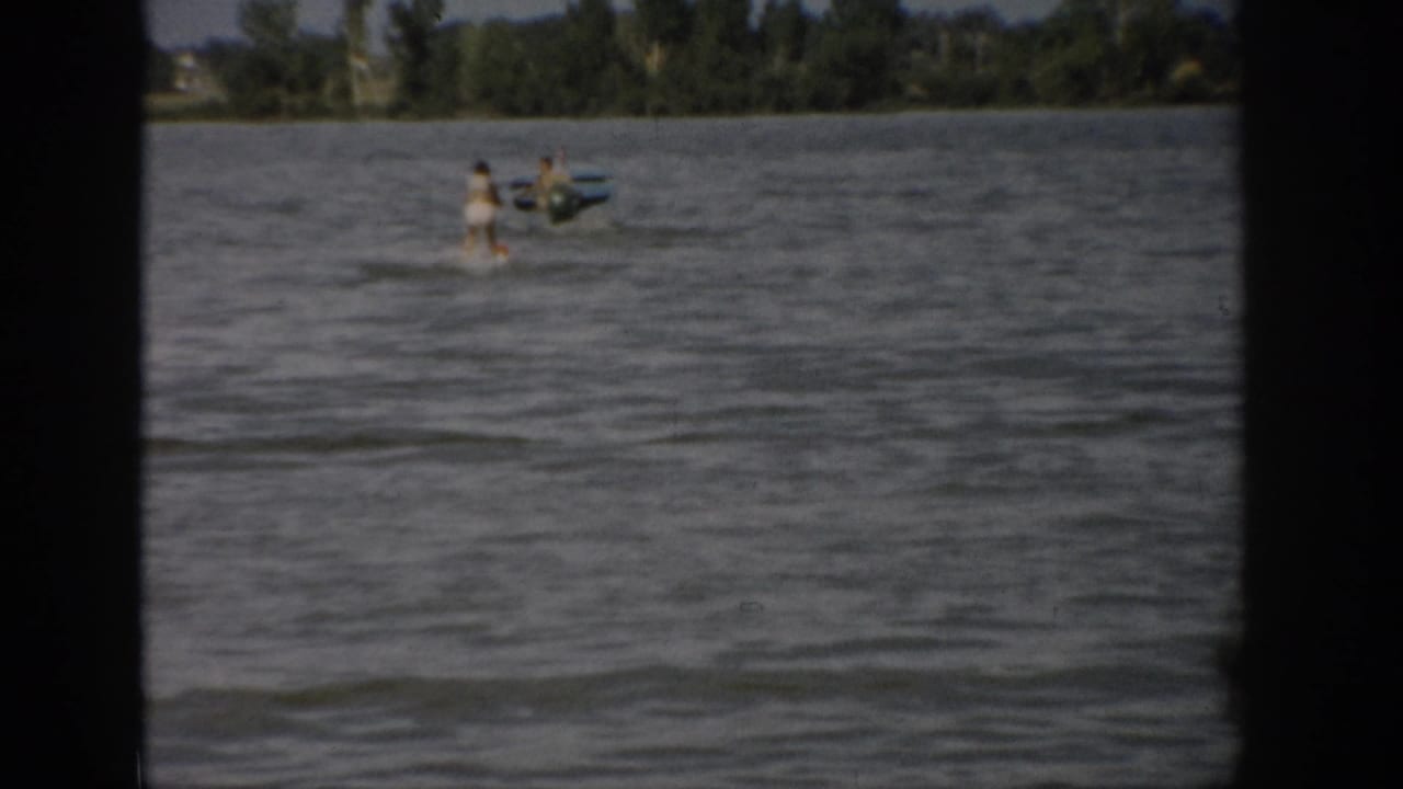 1959 Colorado home-movie frame showing a distant woman water skiing near a small motorboat on a windy lake.